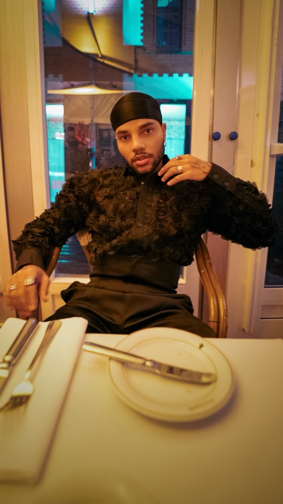 A man in a black outfit and headscarf sits at a table set for dinner. He is wearing a textured black jacket and pants, with rings on his fingers. The setting appears to be a restaurant with a window view.