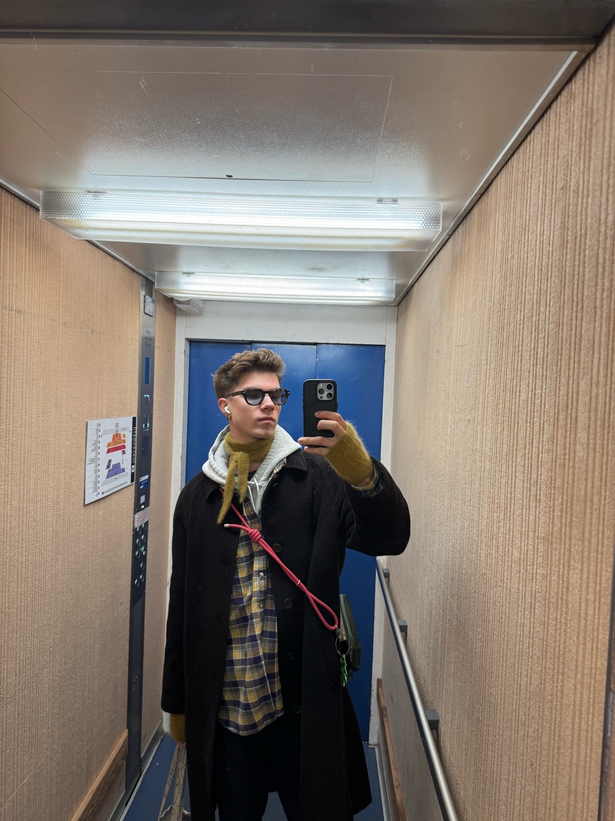 A man takes a selfie in an elevator, showcasing his outfit. He wears glasses, a coat, and a stylish shirt.