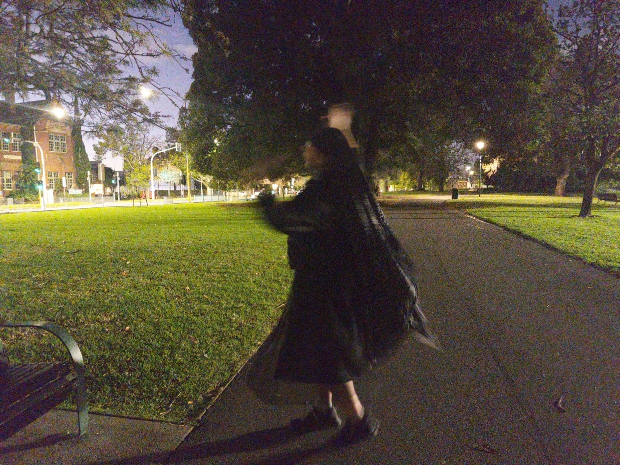 A person in a long black coat dances in a park at night. The grass is green and there are trees in the background.