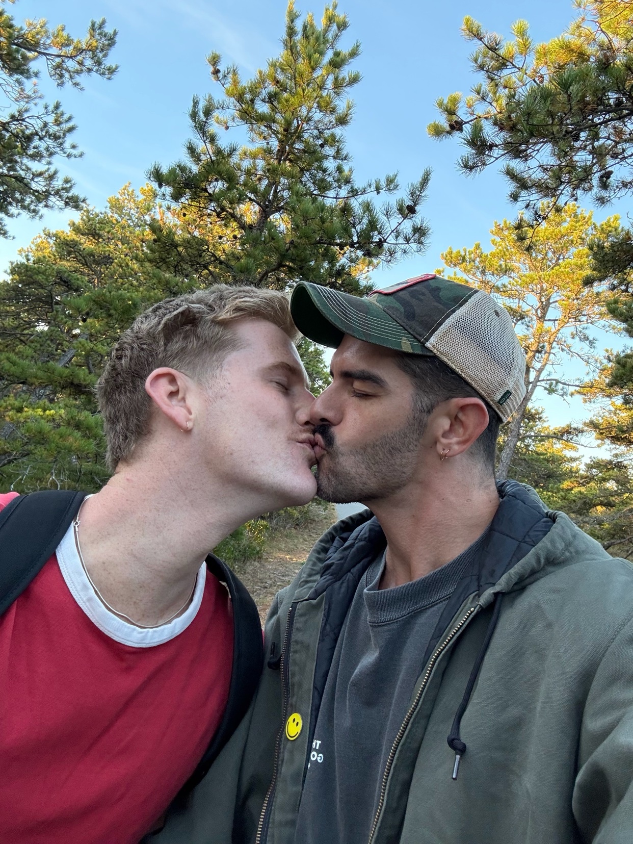 A couple shares a kiss outdoors. The men are embracing in front of a backdrop of trees.