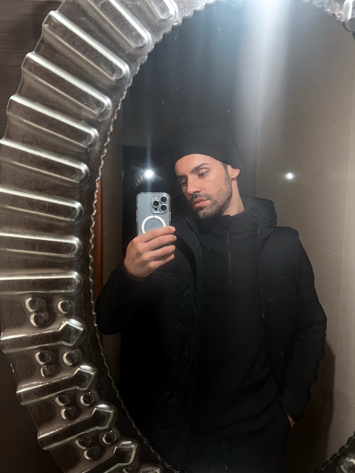A man takes a mirror selfie, showcasing his winter style. He's wearing a black beanie and jacket.