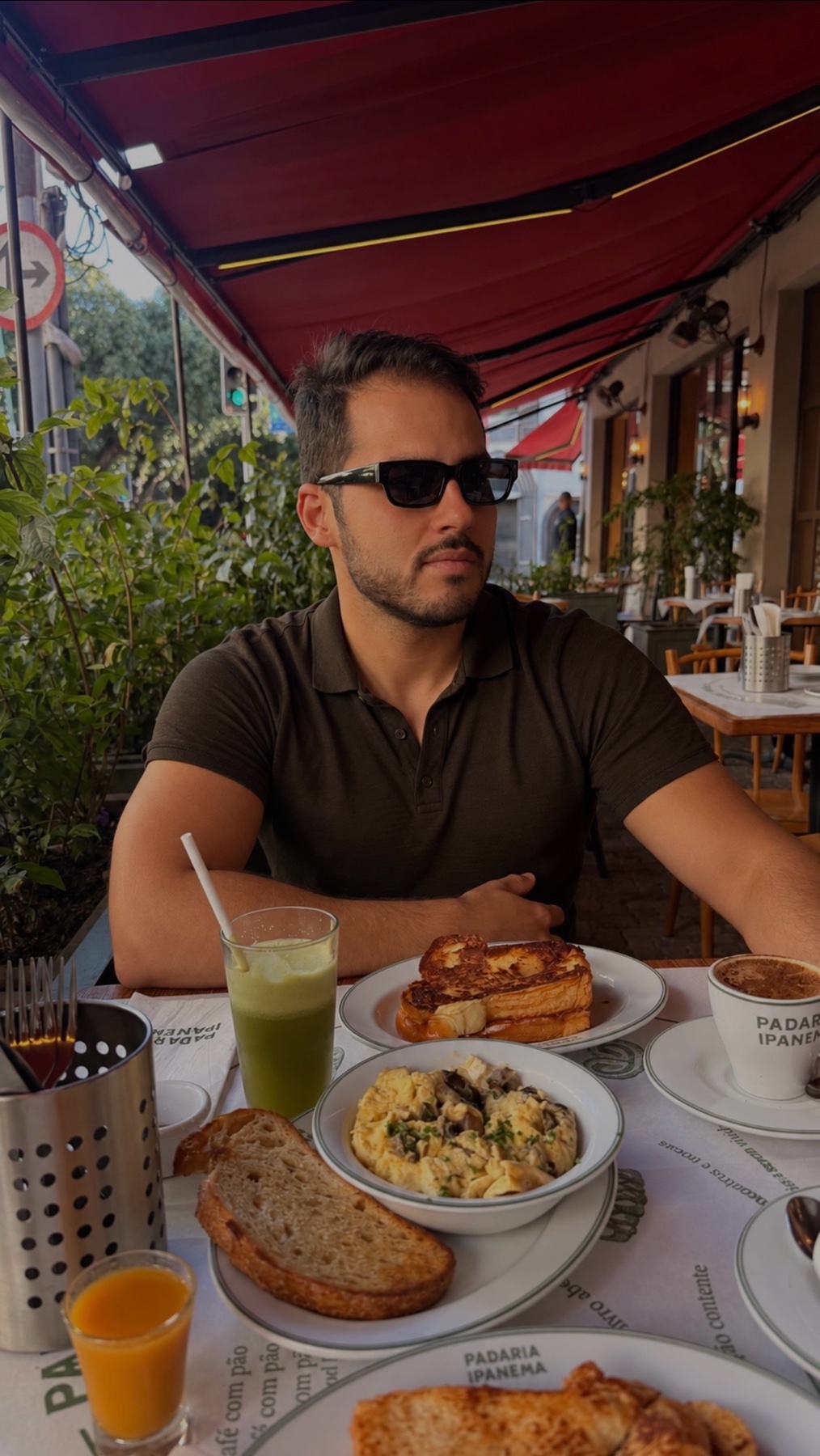 A man wearing sunglasses sits at a table laden with a variety of breakfast foods, including french toast, scrambled eggs, bread, juice, and coffee. He is seated outdoors at a restaurant with red awnings overhead.