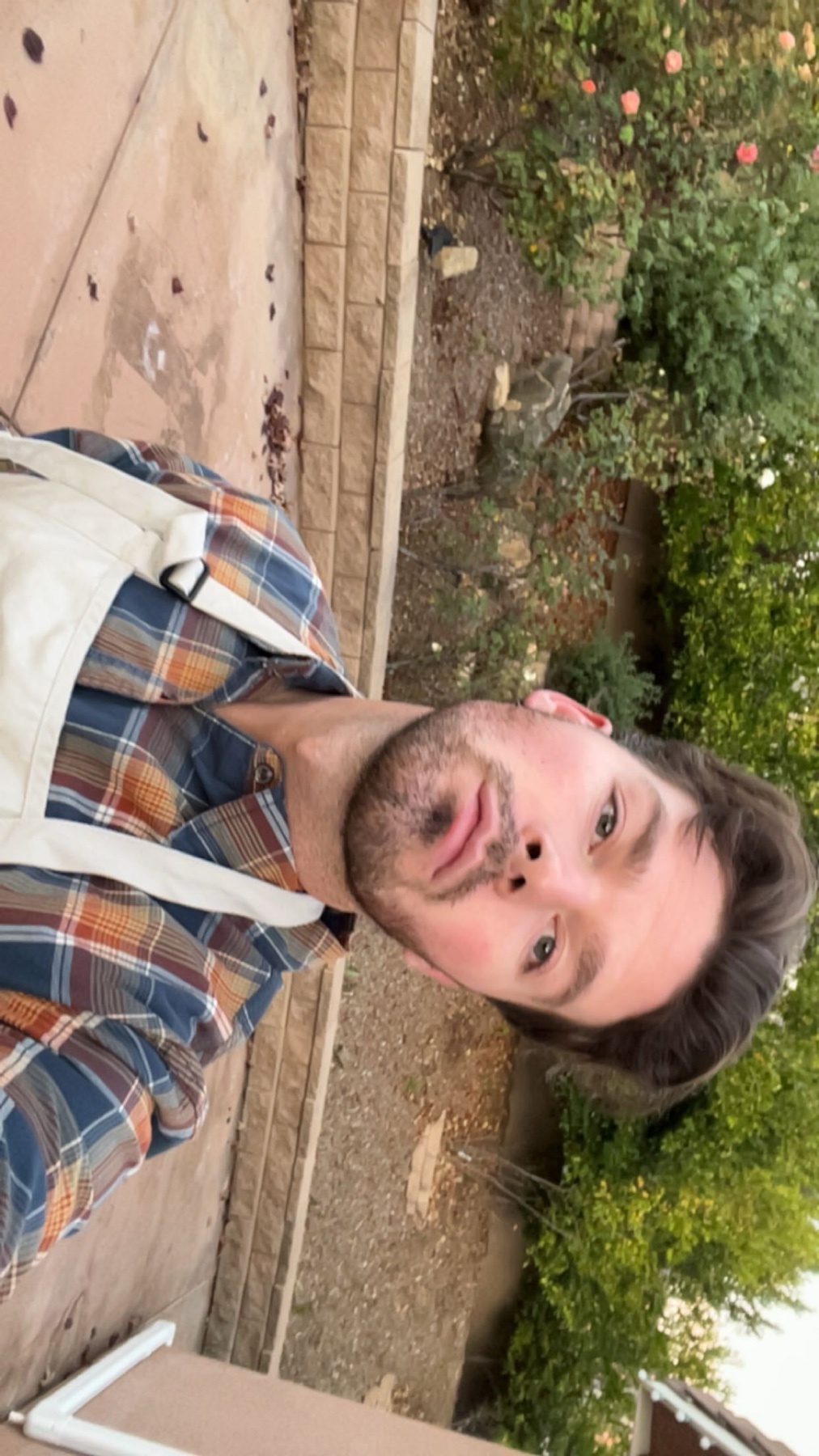 A man takes a selfie while waiting for a kitchen to open. He is wearing a plaid shirt and an apron, standing outside near a garden.
