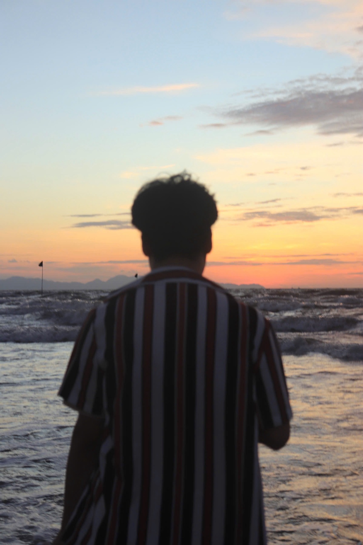 A person stands on a beach, facing the ocean as the sun rises or sets. The sky is painted with warm colors, and waves crash on the shore.