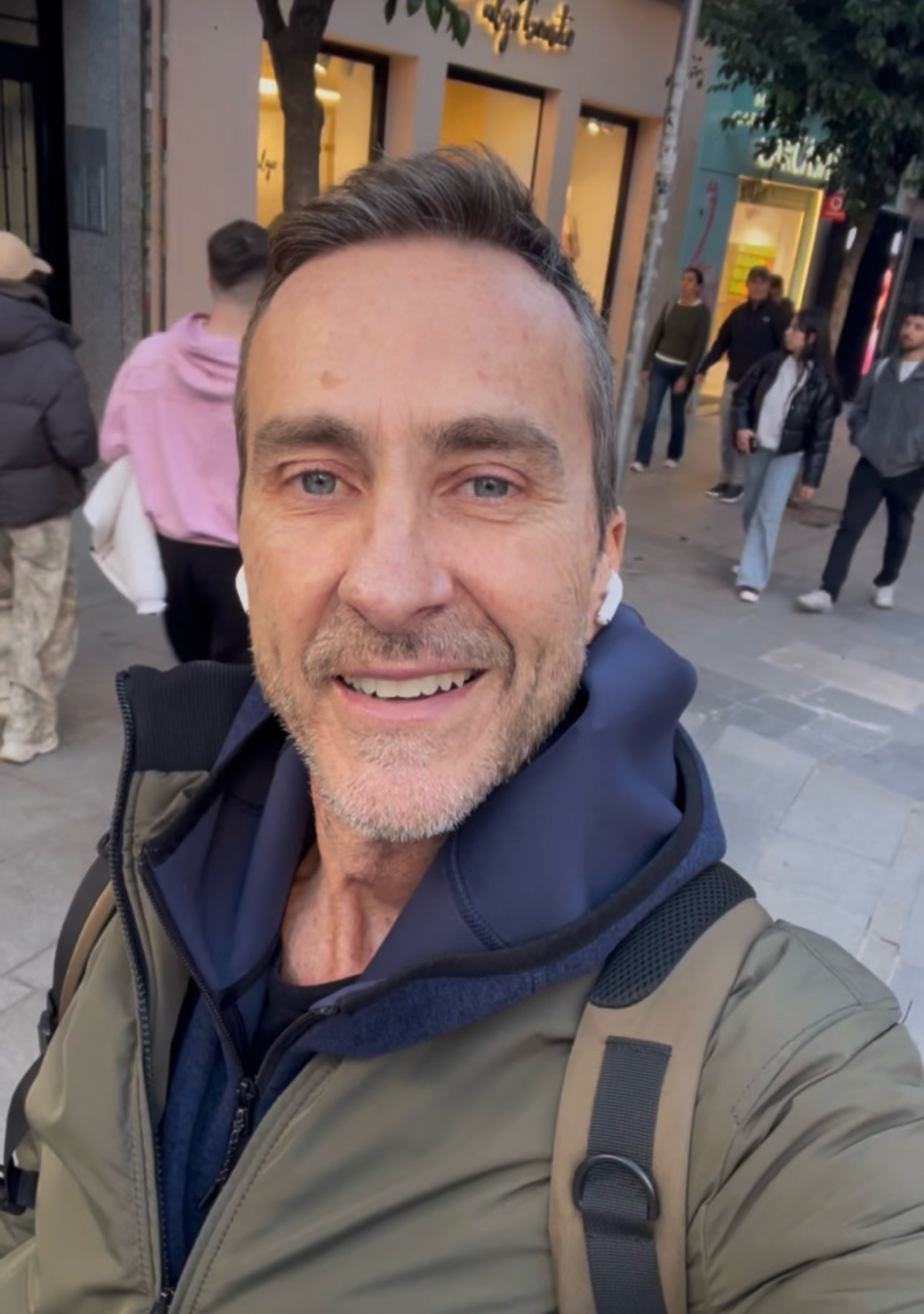 A man takes a selfie on a city street. He is smiling and wearing a casual outfit.