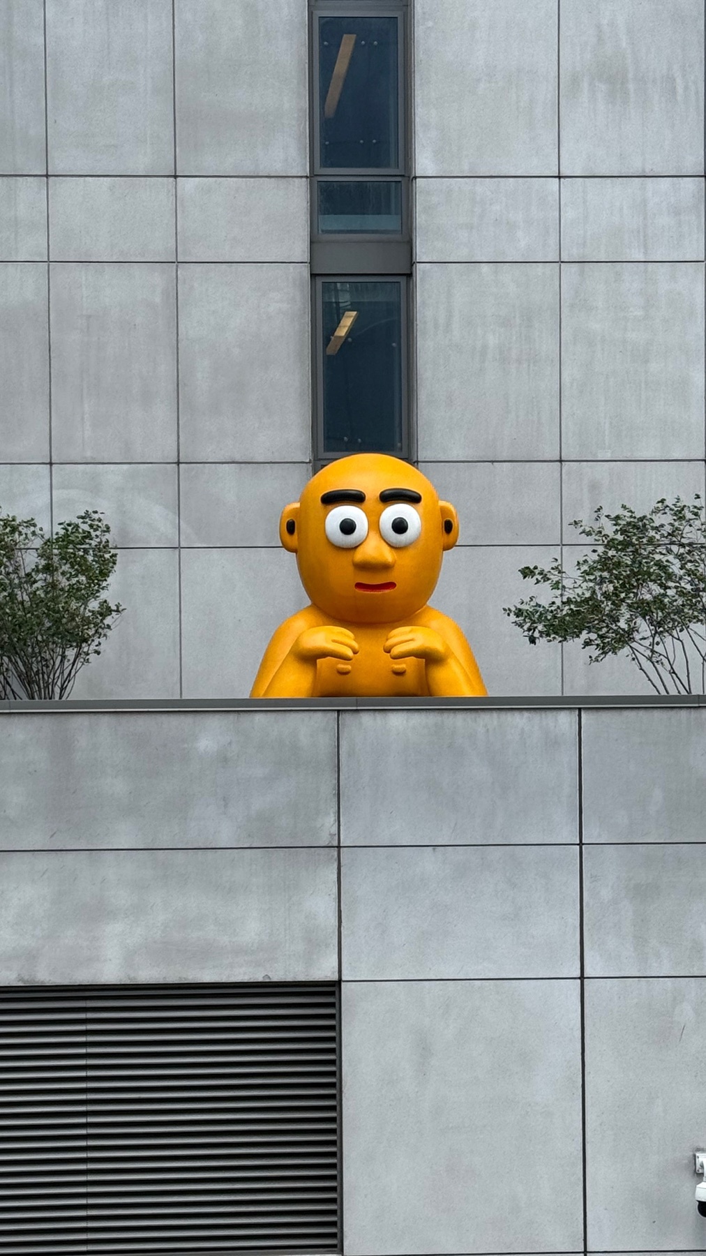 A large, yellow, cartoonish sculpture with wide eyes and a small mouth peers over a modern building. The sculpture is positioned between two small trees, adding a whimsical element to the urban landscape.
