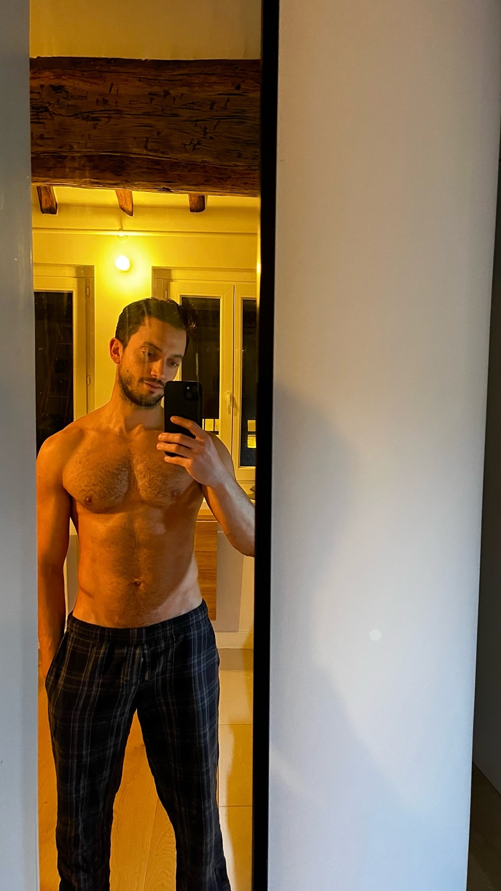 A man takes a shirtless mirror selfie, showcasing his physique. He is wearing pajama pants and has dark hair and stubble.