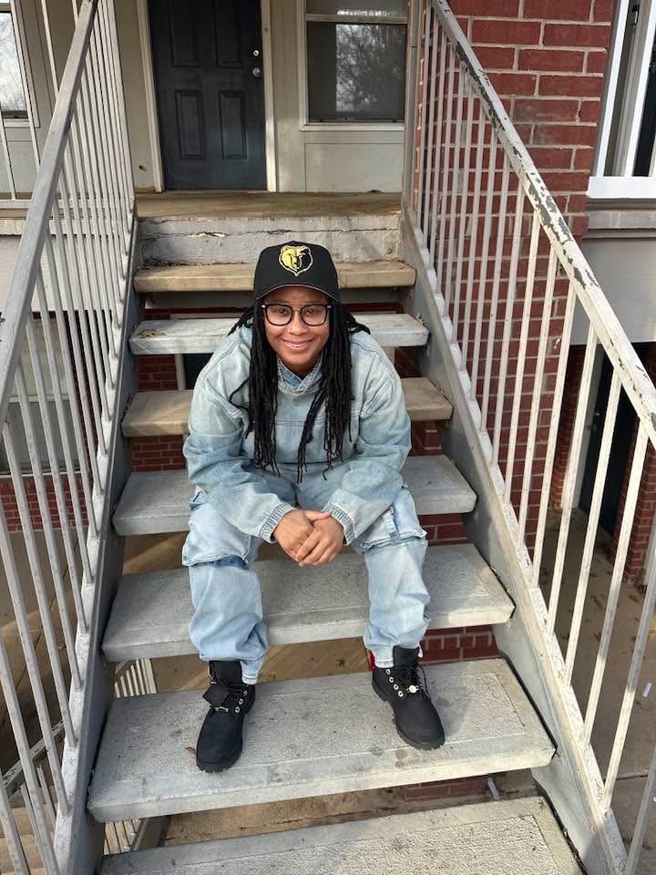 A woman with dreadlocks and glasses sits on outdoor stairs wearing a denim jacket and pants, a baseball cap, and boots.