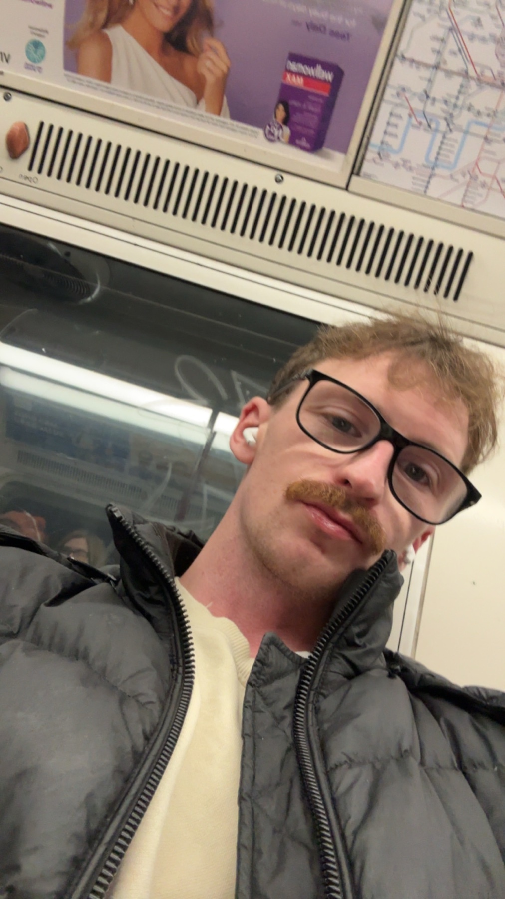 A man with glasses and a mustache takes a selfie on the London Underground. The photo captures a moment of daily life in the city.
