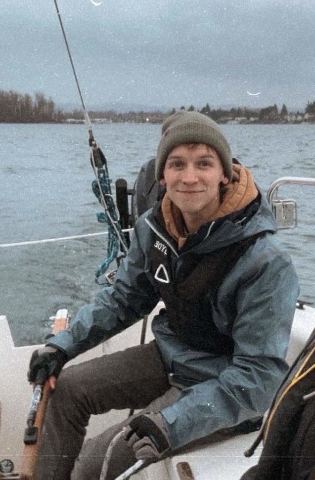 A young man with a beanie and a life vest smiles while sitting on a sailboat. He is wearing a blue jacket and dark pants.