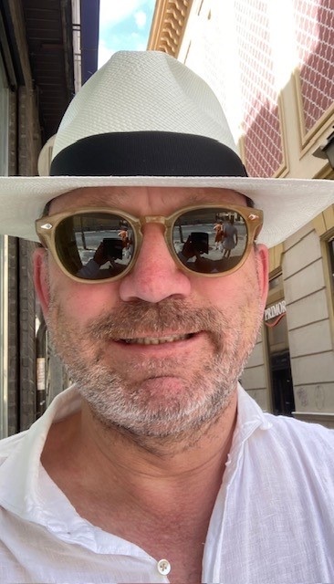 A man wearing a white fedora and sunglasses smiles at the camera. He is wearing a white collared shirt.