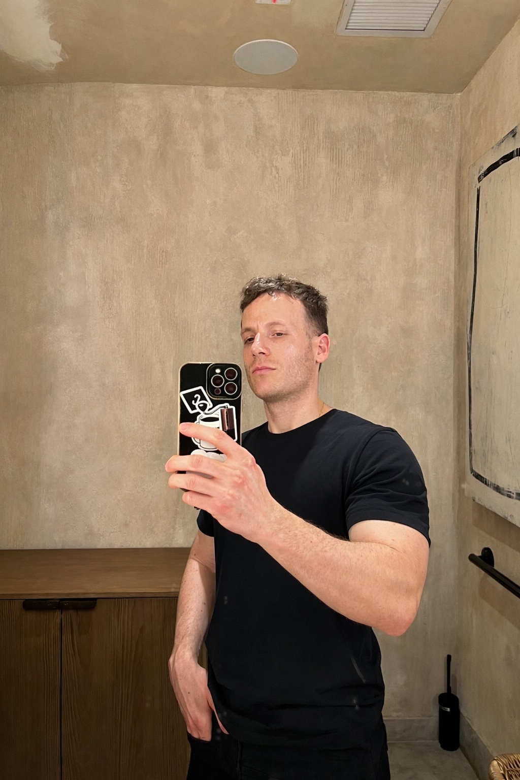 A man takes a mirror selfie in a bathroom. He is wearing a black t-shirt.