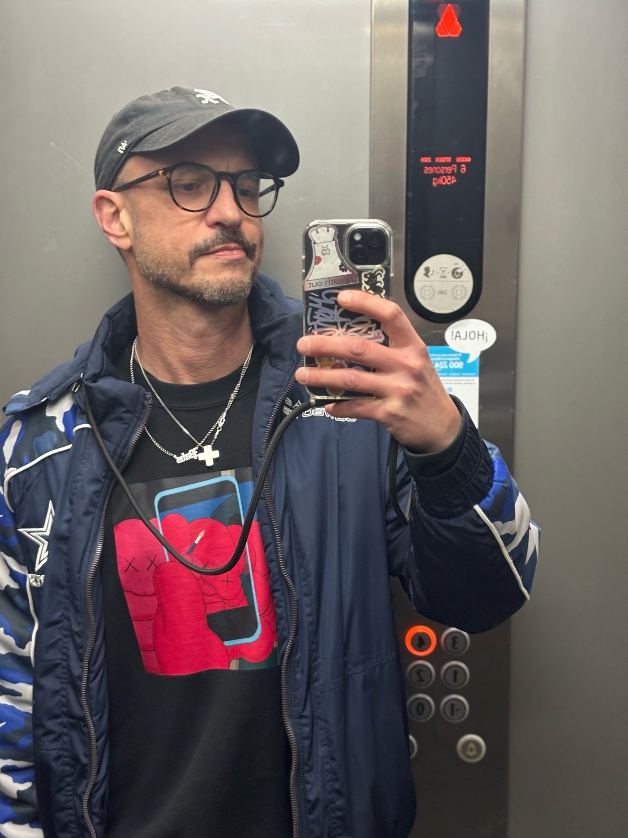 A man in a baseball cap and glasses takes a selfie in an elevator. He is wearing a dark jacket over a graphic t-shirt and multiple necklaces.