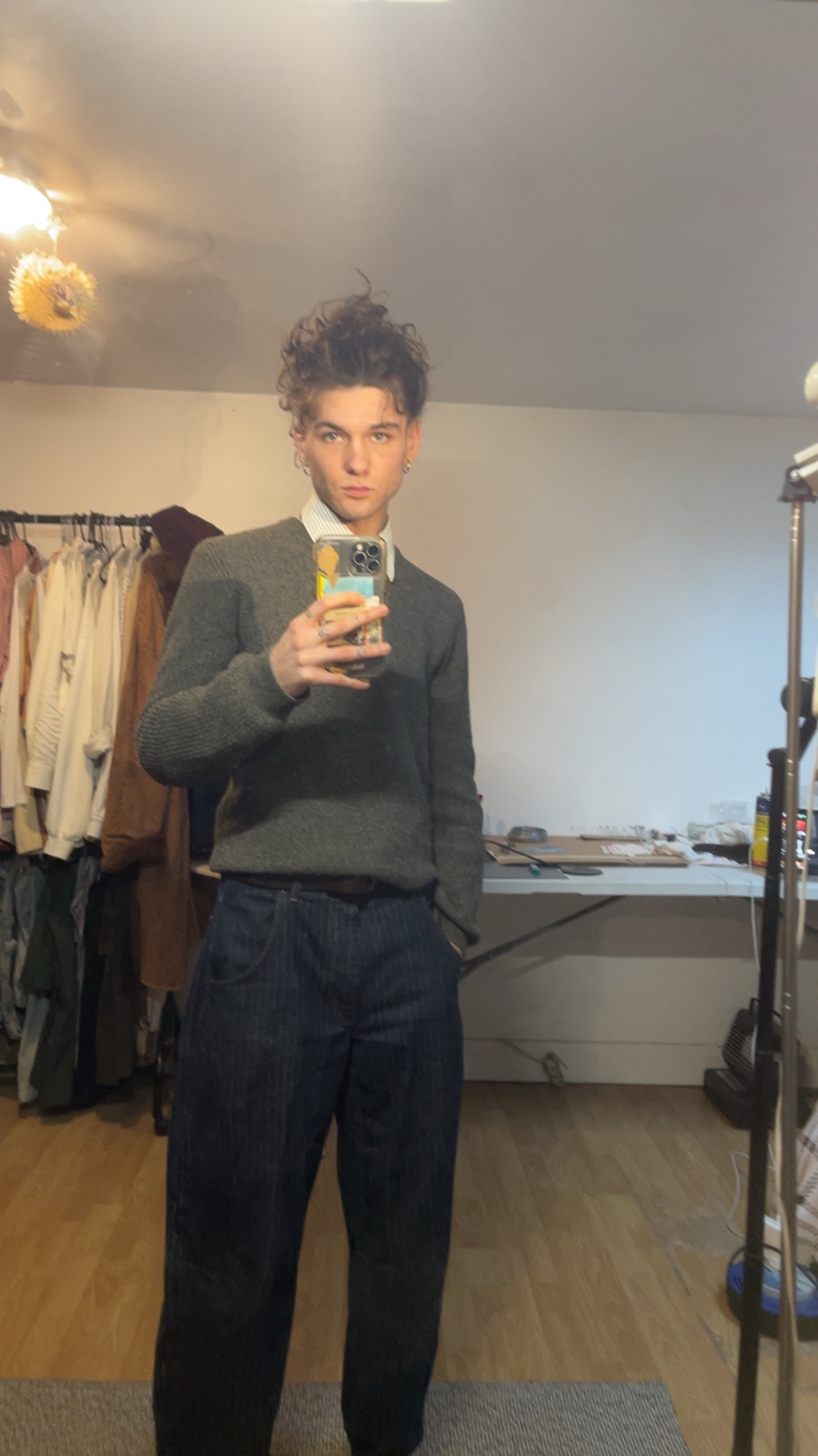 A man takes a mirror selfie, showcasing his outfit. He's wearing a sweater and jeans, with curly hair.