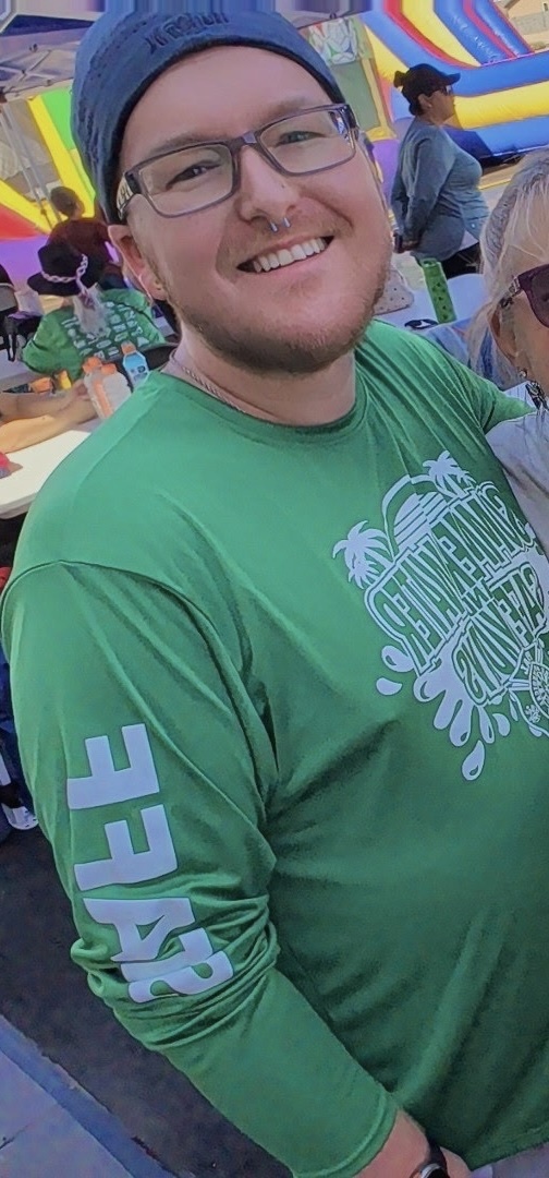 A man wearing glasses, a beanie, and a green long-sleeved shirt smiles at the camera. In the background, an inflatable slide and other people are visible.