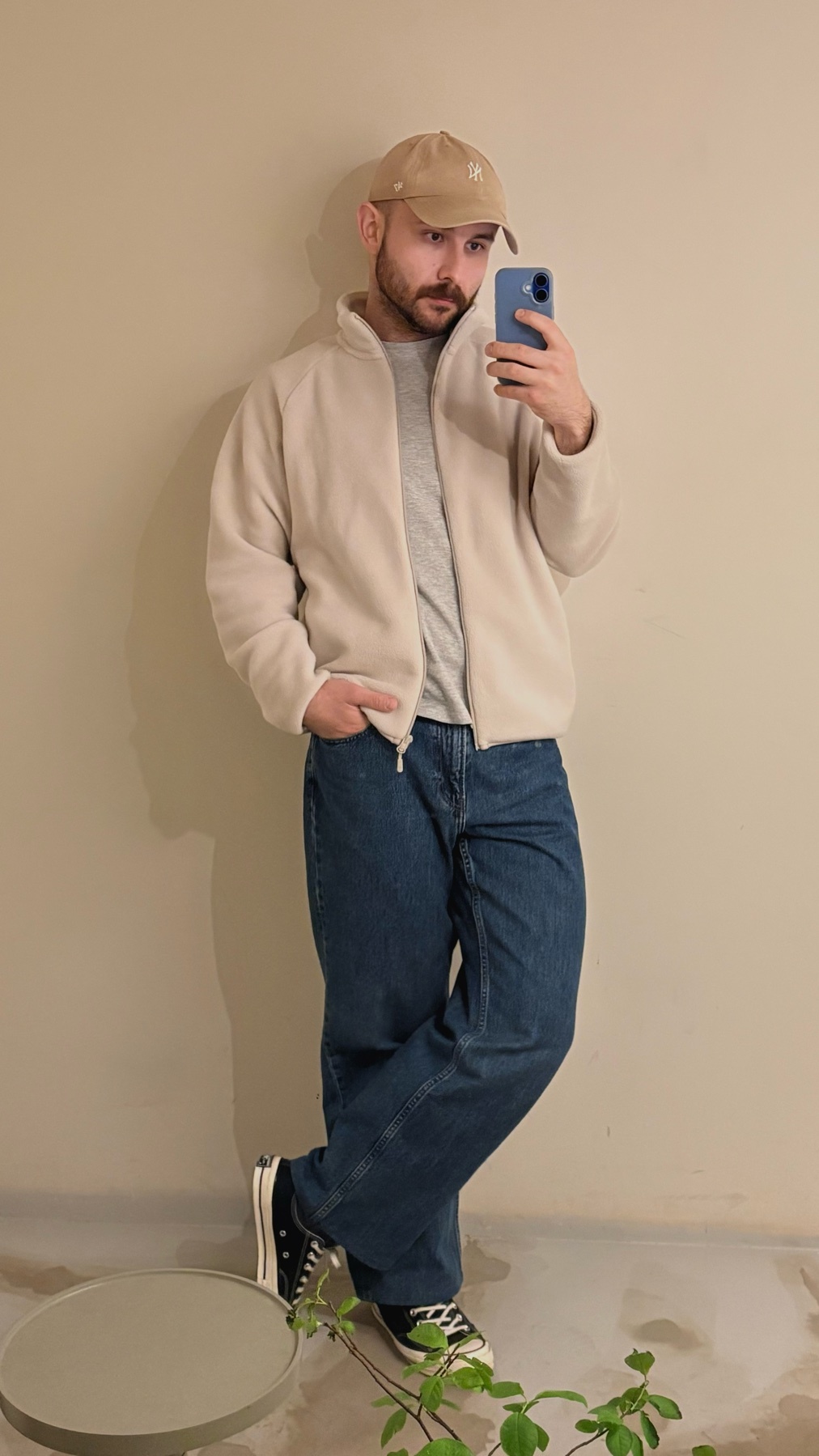 A man in a tan baseball cap, grey t-shirt, cream fleece jacket, and blue jeans poses for a selfie against a neutral wall. He is wearing black sneakers and has his hand in his pocket.