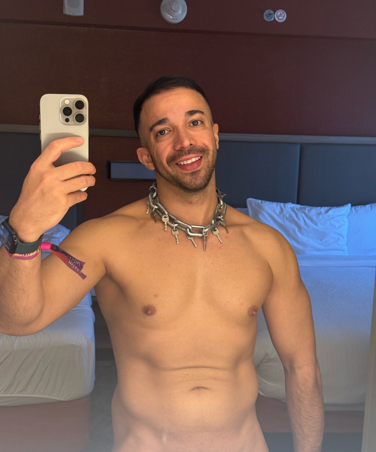 A shirtless man with short dark hair and a spiked collar smiles at the camera while taking a selfie in a hotel room.