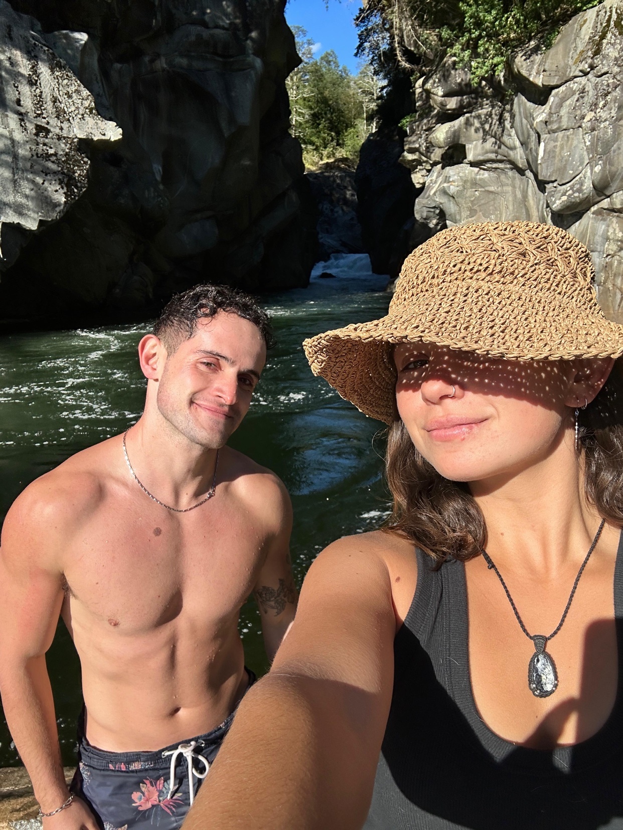 A shirtless man with a muscular physique and a woman wearing a hat take a selfie by a river. They are surrounded by rocky cliffs and lush greenery, suggesting an outdoor adventure.