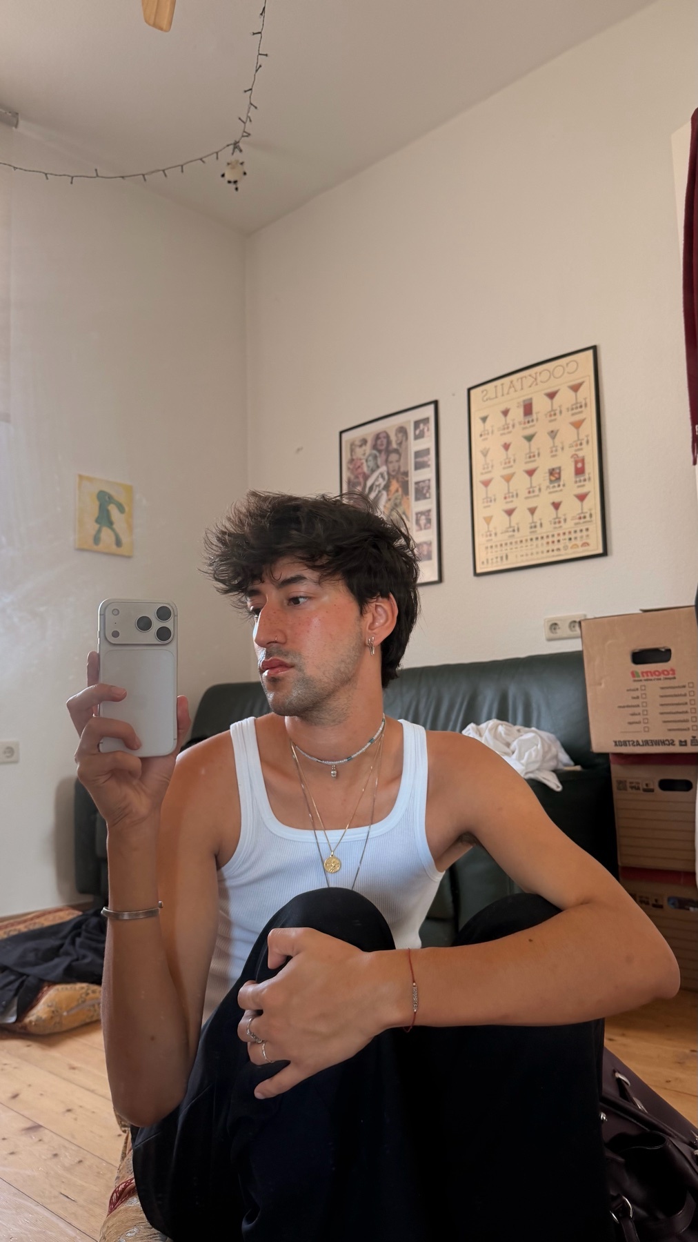 A young man takes a mirror selfie in a room, wearing a white tank top and dark pants. He has dark, messy hair and is adorned with necklaces and bracelets.