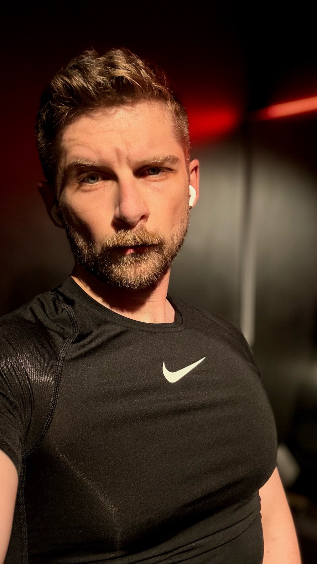 A man in a black workout shirt takes a selfie in a gym. The photo is lit with warm light.
