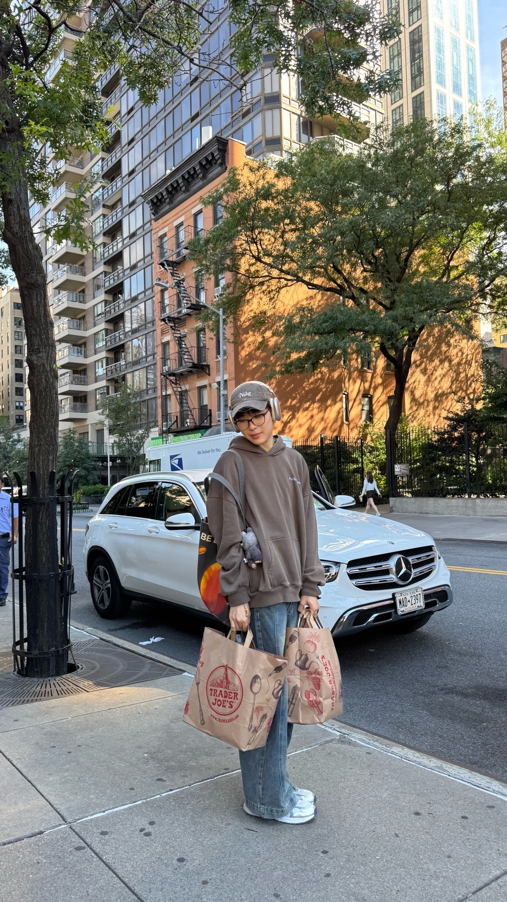 A woman stands on a sidewalk in a city, holding two grocery bags from Trader Joe's. She is wearing a brown hoodie, baggy jeans, and a baseball cap, embodying a casual street style.