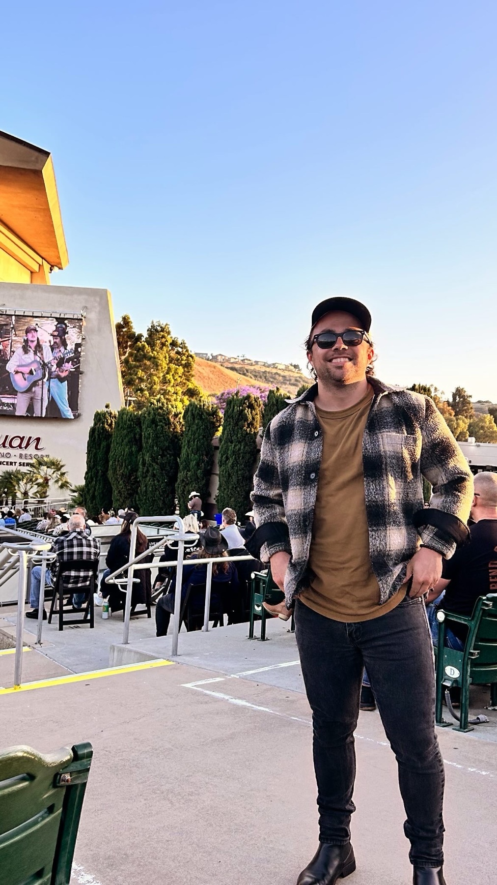 A man poses for a photo at an outdoor concert venue. He is wearing a plaid shirt, jeans, and sunglasses.