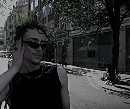 A man with curly dark hair, stubble, and a mustache poses for a street style photo. He wears sunglasses, a chain necklace, and a ring on his finger.