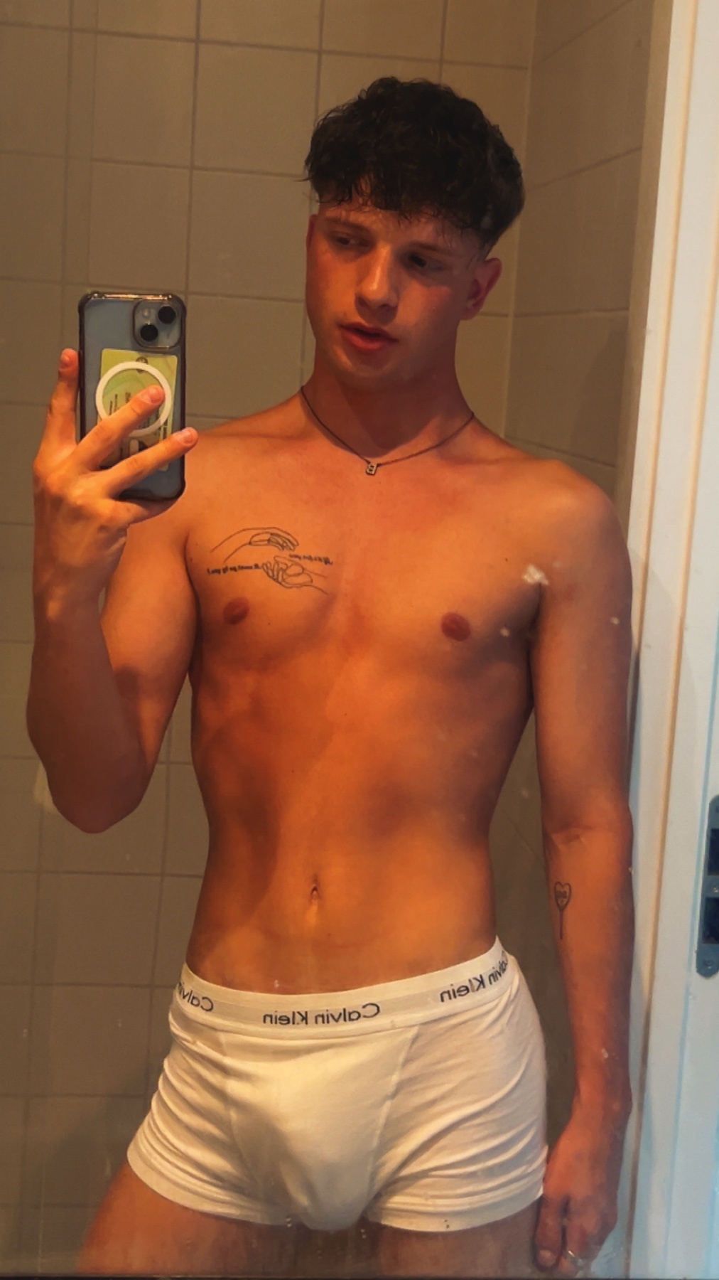 A young man with curly dark hair poses for a mirror selfie in white Calvin Klein underwear. He has tattoos on his chest and arm.