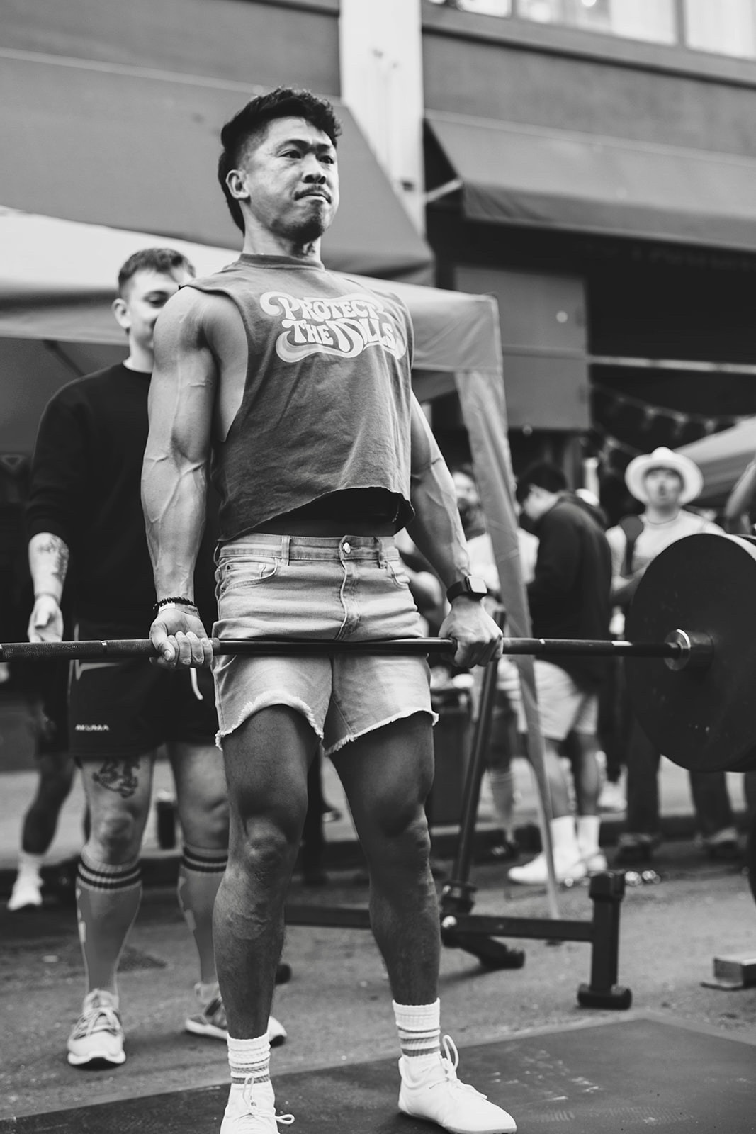 A man lifts a barbell in an outdoor weightlifting competition. He is muscular and focused, with a spotter behind him.