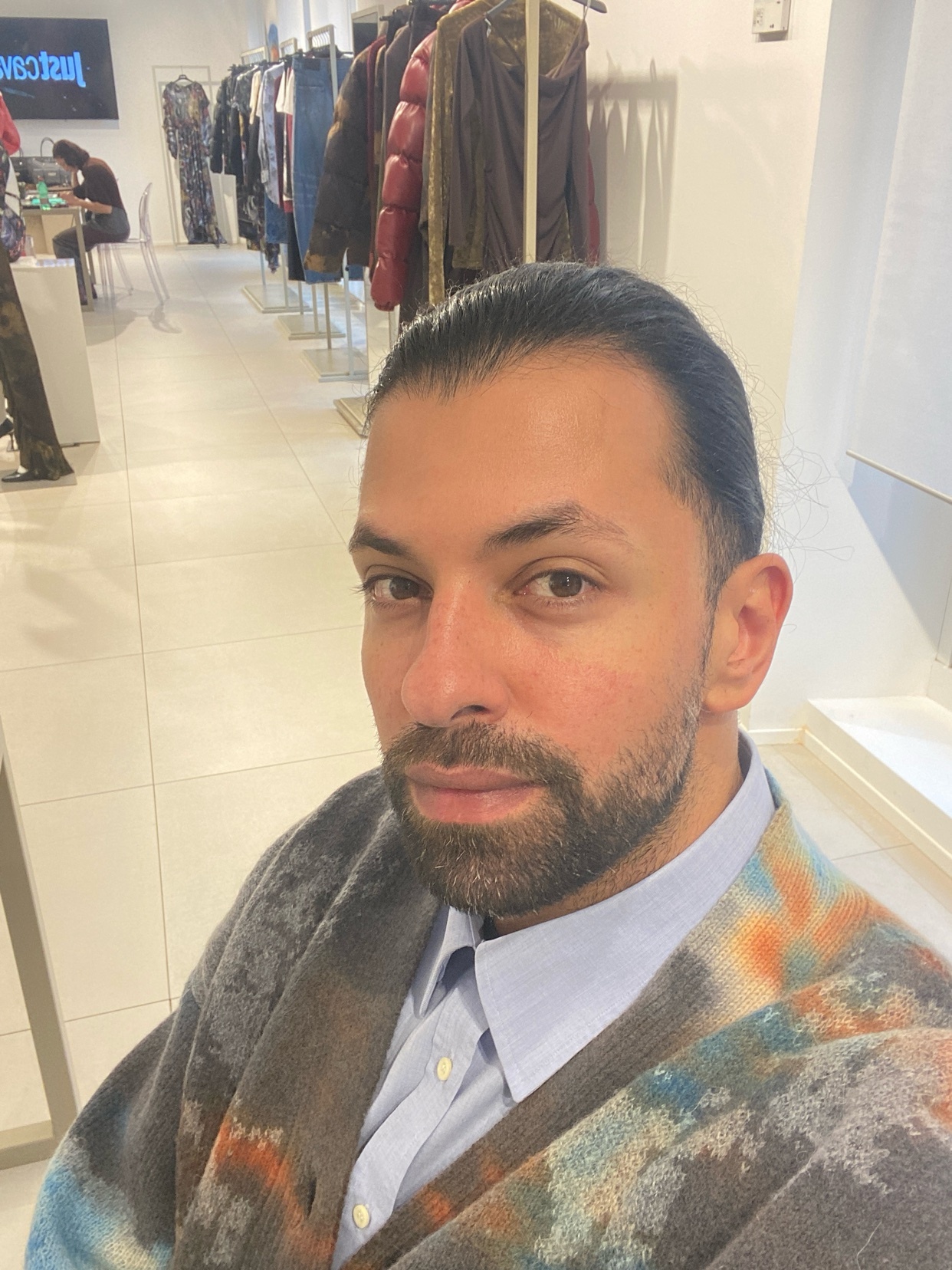 A man takes a selfie in a clothing store. He is wearing a patterned cardigan and a collared shirt.