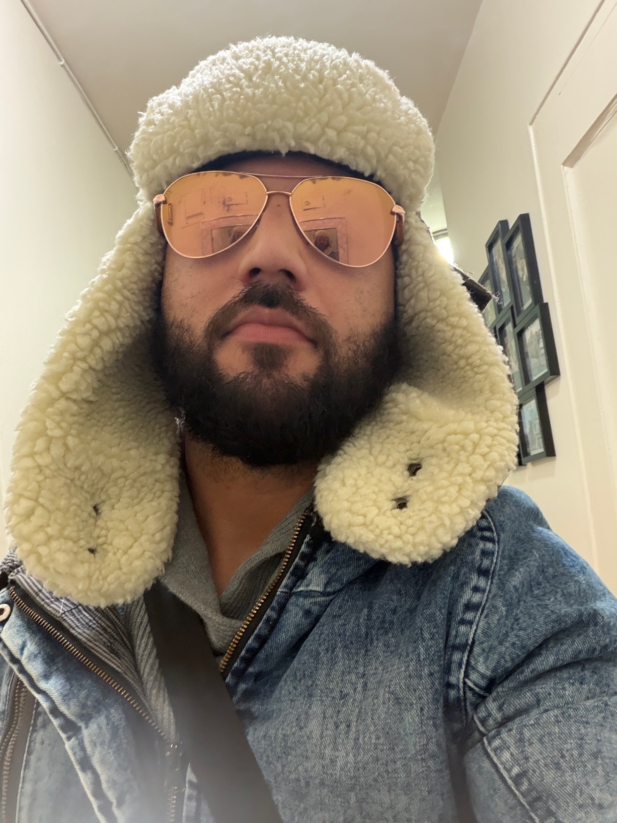 A man wearing a winter hat and sunglasses takes a selfie. He is wearing a denim jacket.