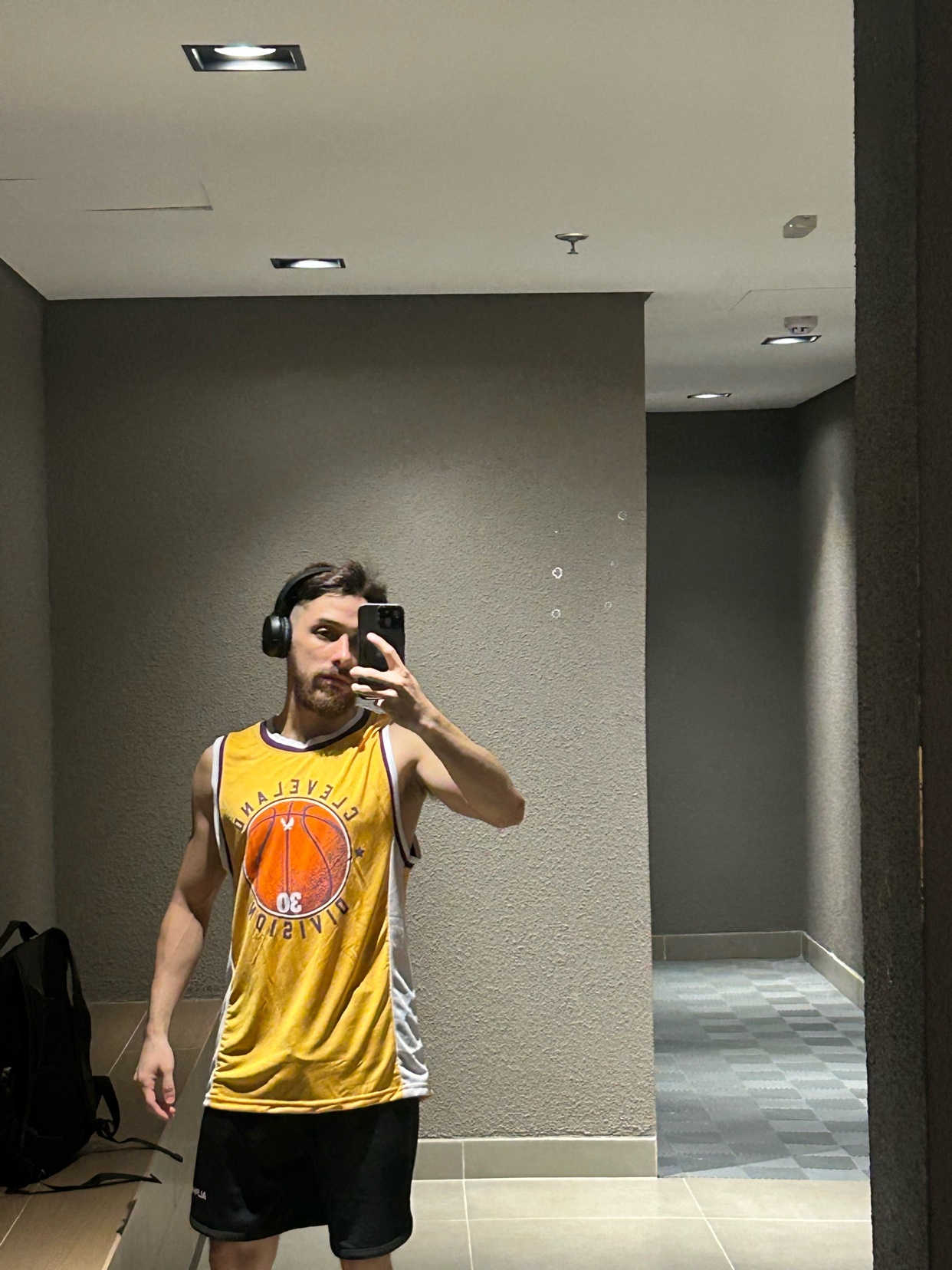 A man takes a mirror selfie in a gym, wearing a basketball jersey and headphones. He appears to be working out or preparing for a workout.