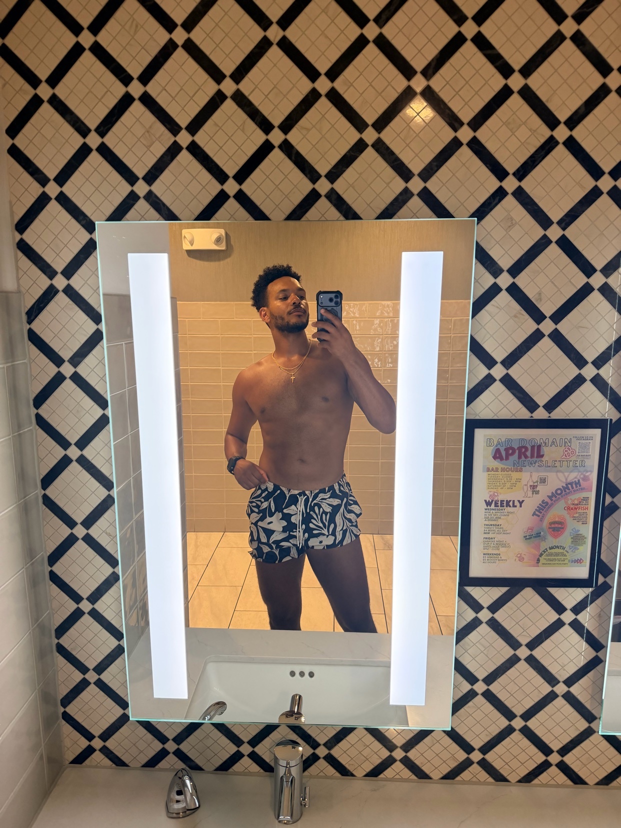A man takes a mirror selfie in a bathroom, wearing patterned swim trunks. The background features a black and white diamond tile pattern on the wall.