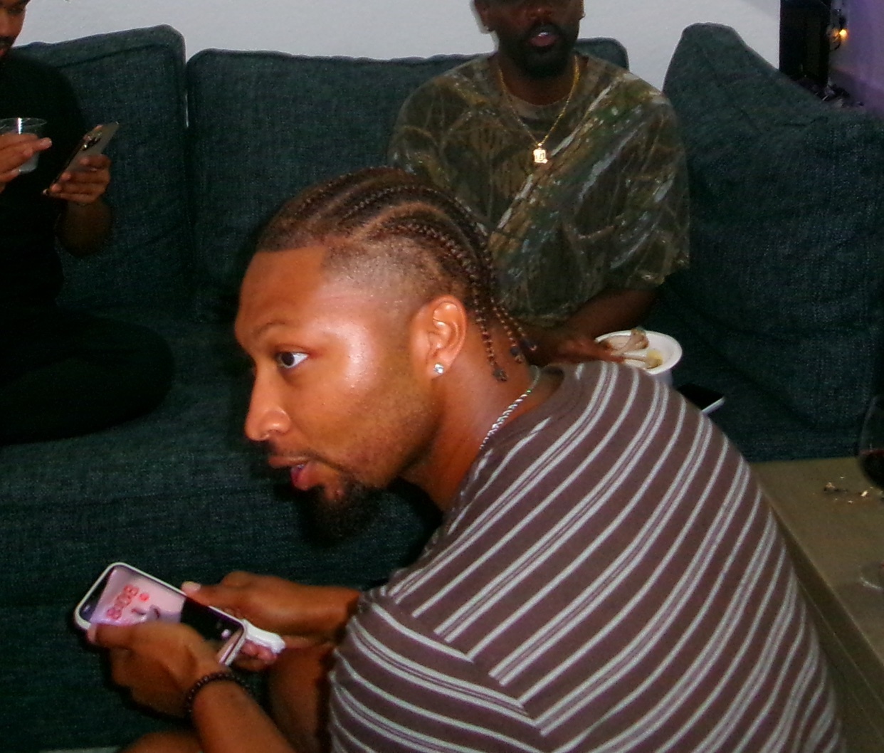 A man with braided hair is looking to the side while holding a phone. He is wearing a brown and white striped shirt and is seated on a couch with other people in the background.