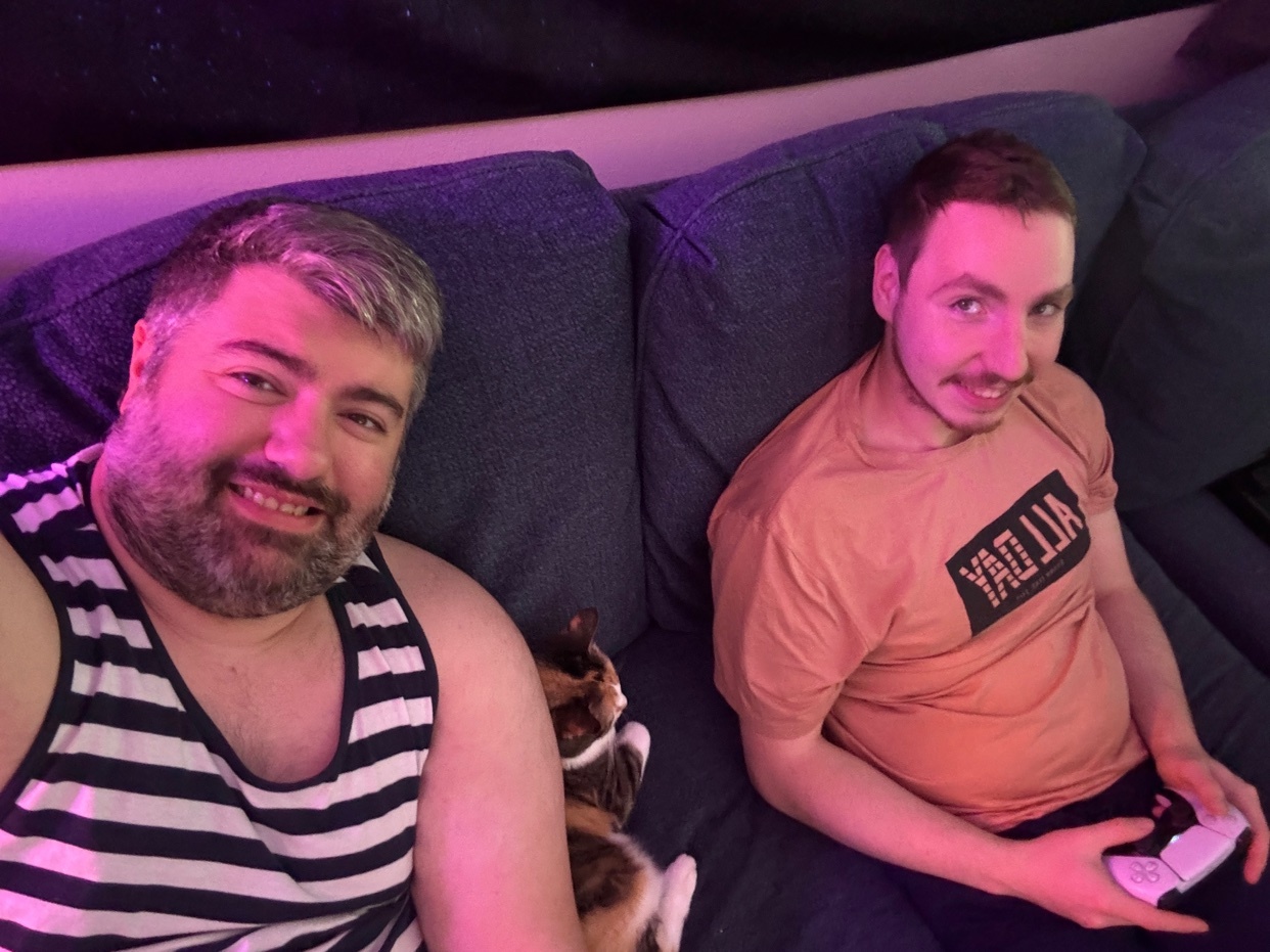 Two men and a cat are relaxing on a couch. The man on the left is wearing a striped tank top and has a beard. The man on the right is wearing a t-shirt and holding a video game controller.