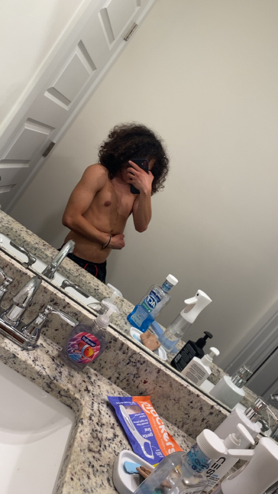 A shirtless man with curly hair takes a mirror selfie in a bathroom, flexing his arm.