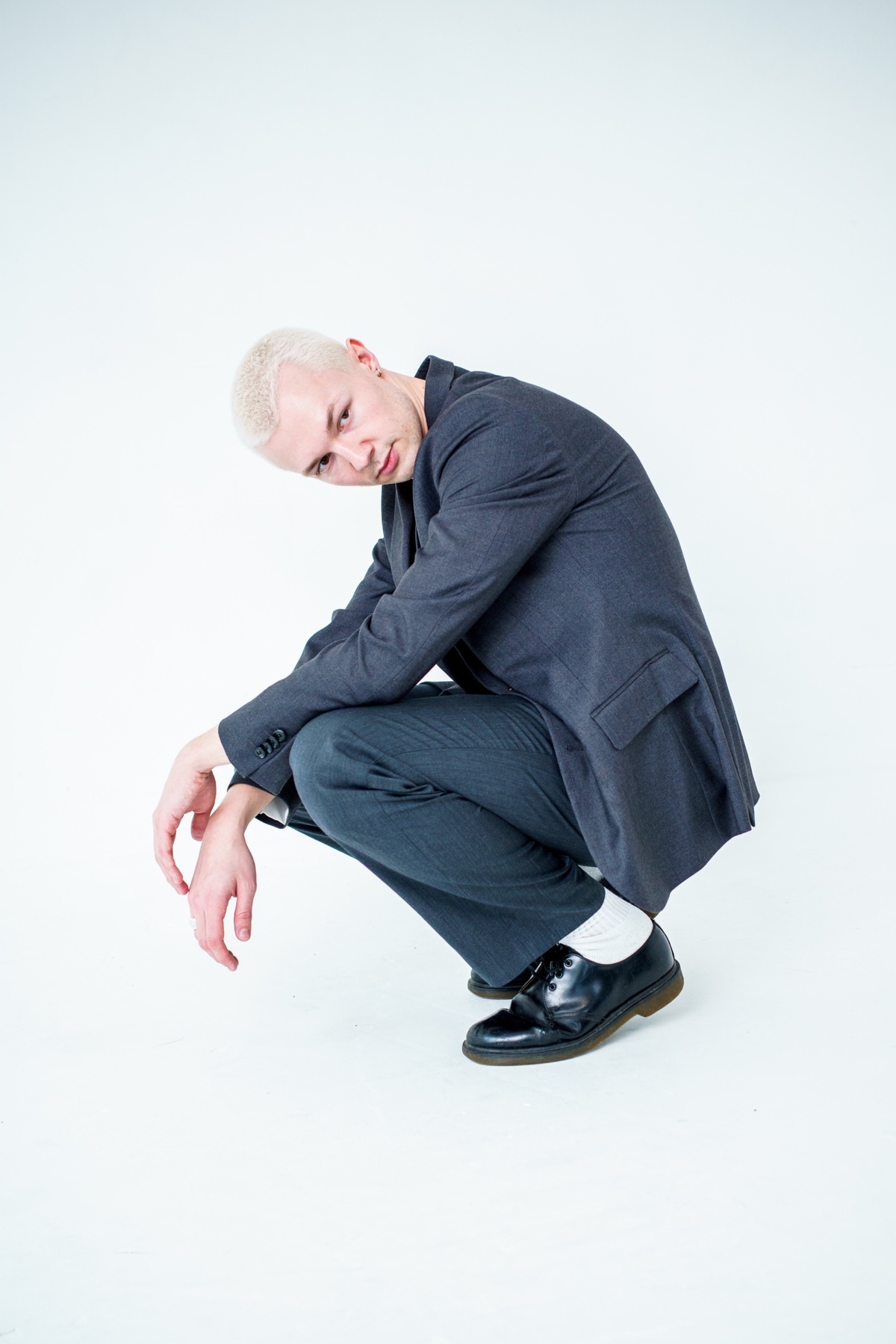 A man in a suit squats in a studio setting. The photo showcases a modern and stylish outfit.