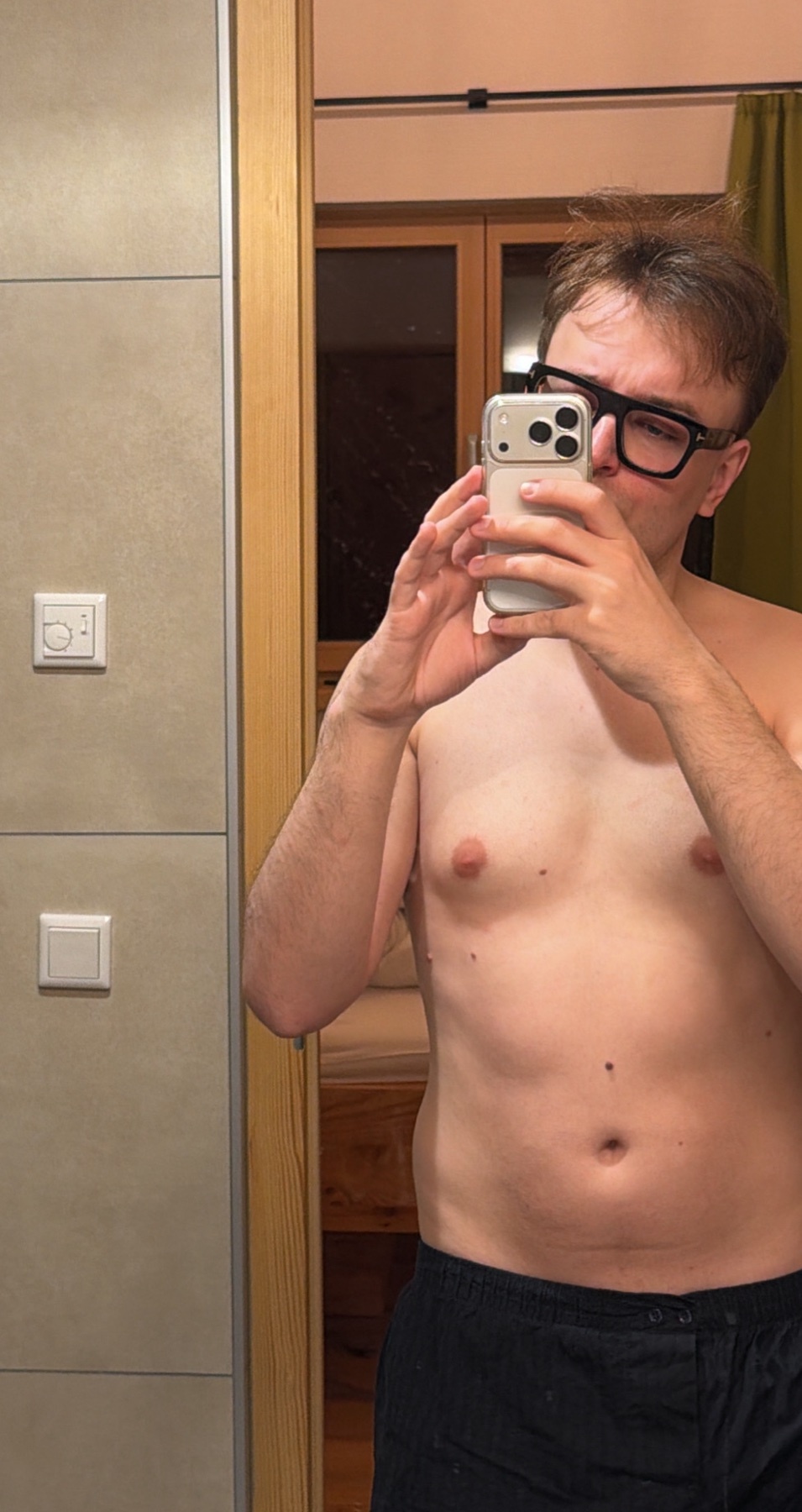 A man in glasses takes a mirror selfie in a bathroom, wearing dark shorts and no shirt.