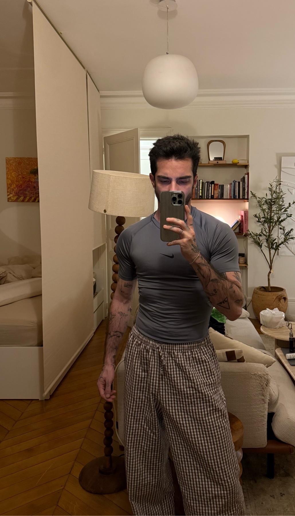 A man takes a mirror selfie in his bedroom. He is shirtless and wearing pants.