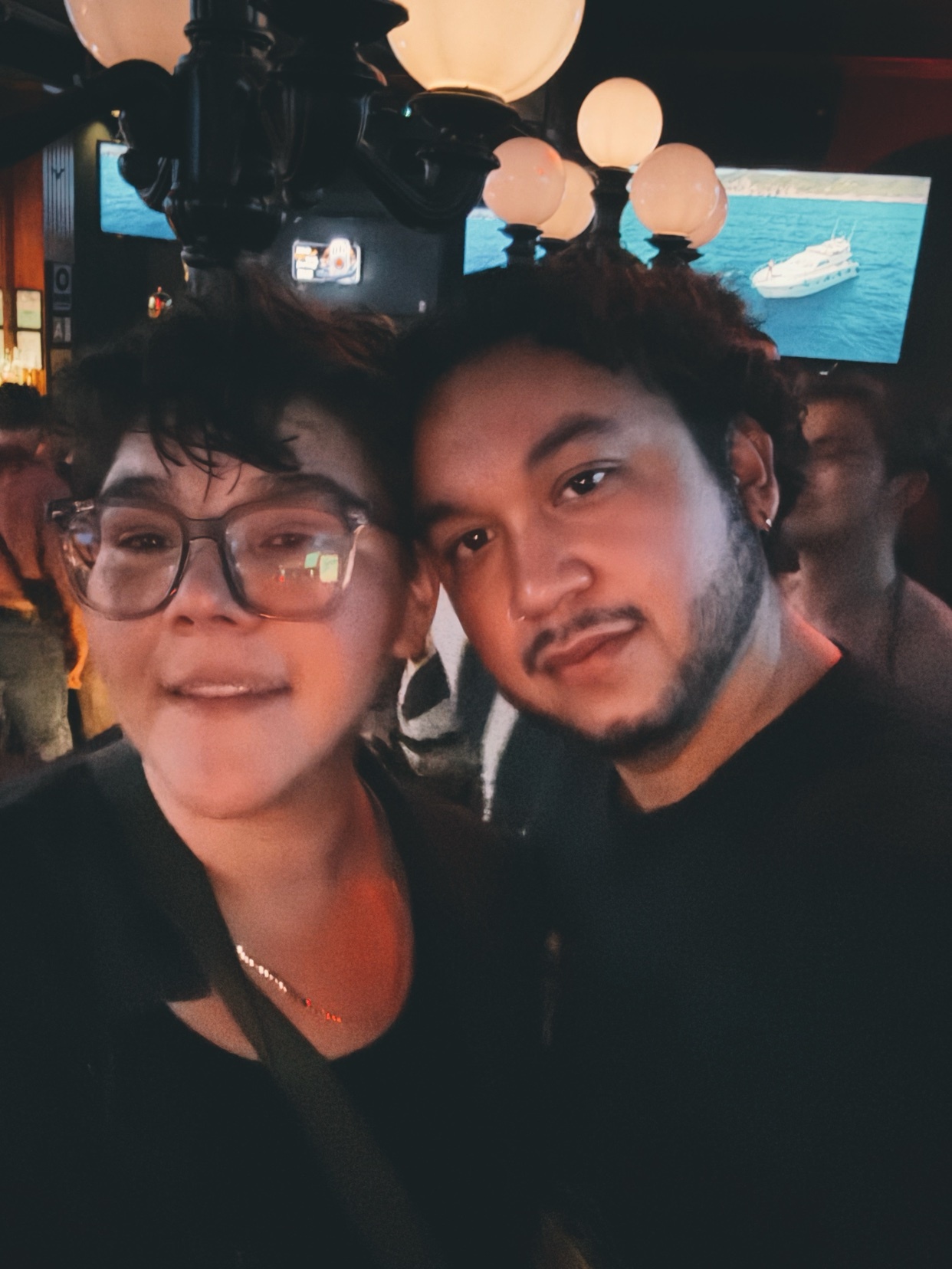 A close-up selfie of two friends, one wearing glasses and the other with a beard, posing together at night.