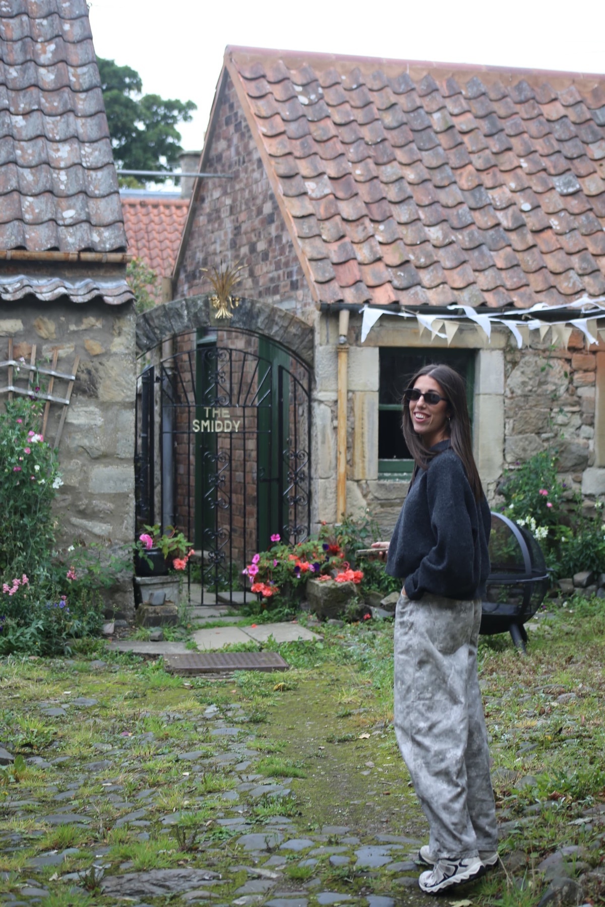 A woman with long dark hair and sunglasses stands in front of a rustic stone building with a tiled roof. She is wearing a dark sweatshirt and patterned pants, with a small garden and flag bunting visible.
