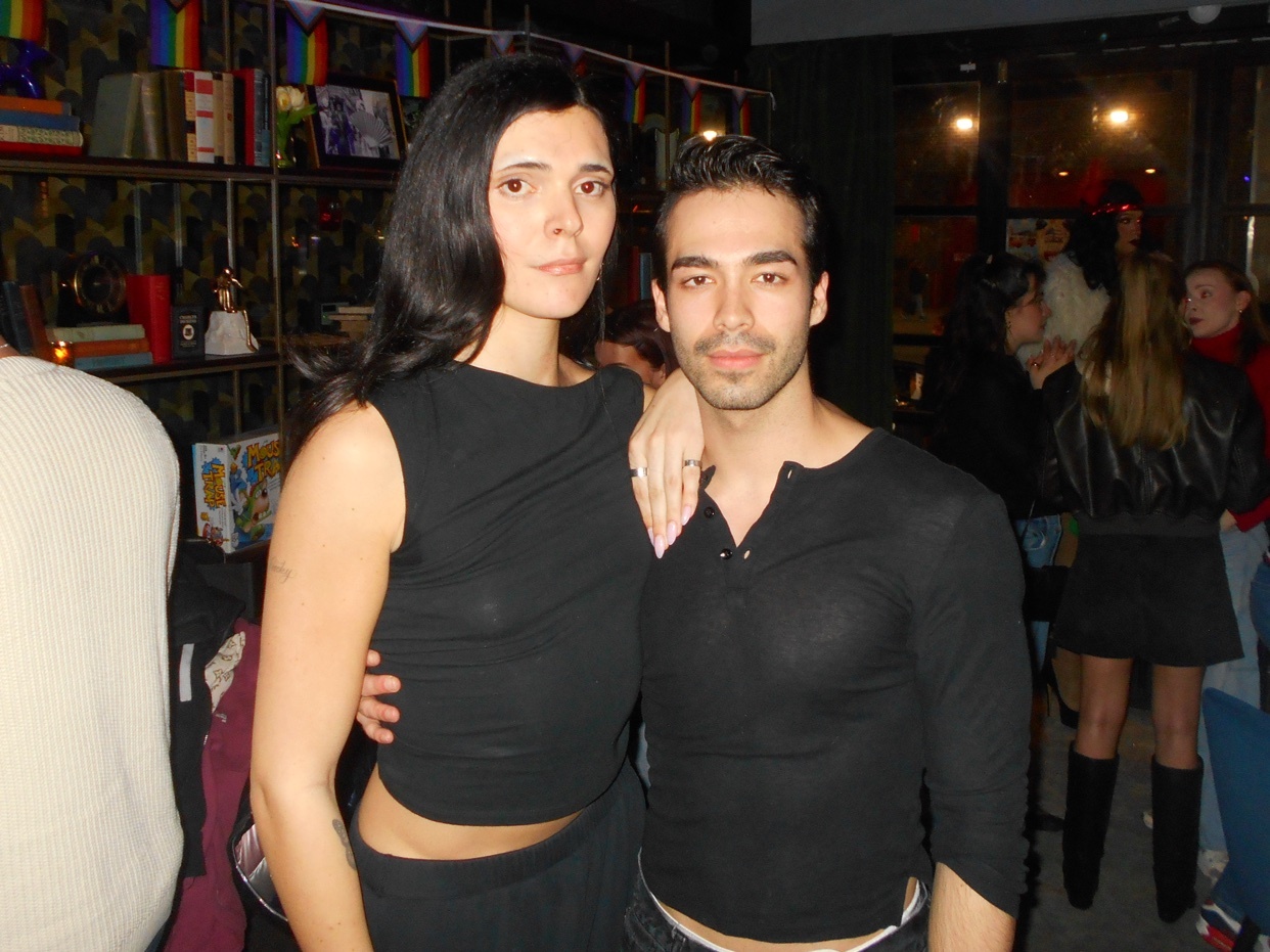A woman and a man pose for a photo in what appears to be a bar or club. The woman has long dark hair and is wearing a black sleeveless top and a dark skirt. The man has short dark hair and is wearing a black long-sleeved shirt. Rainbow flags are visi