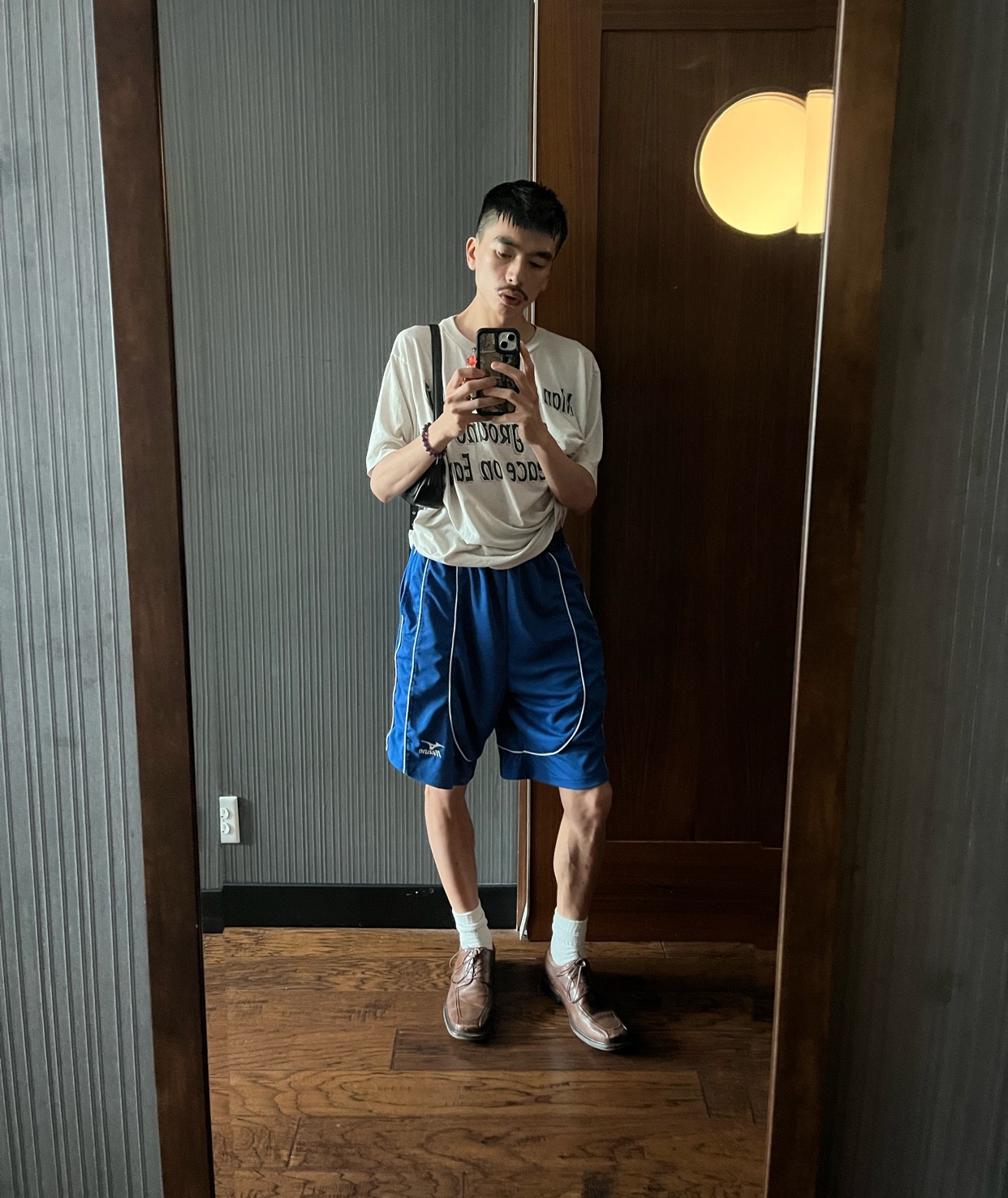 A man takes a mirror selfie, showcasing his outfit. He is wearing a t-shirt, blue shorts, and sneakers.