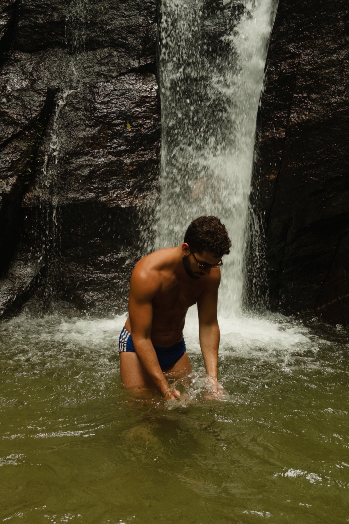 A man stands in a waterfall, shirtless and wearing blue swim trunks. The photo captures a moment of relaxation and adventure in a natural setting.