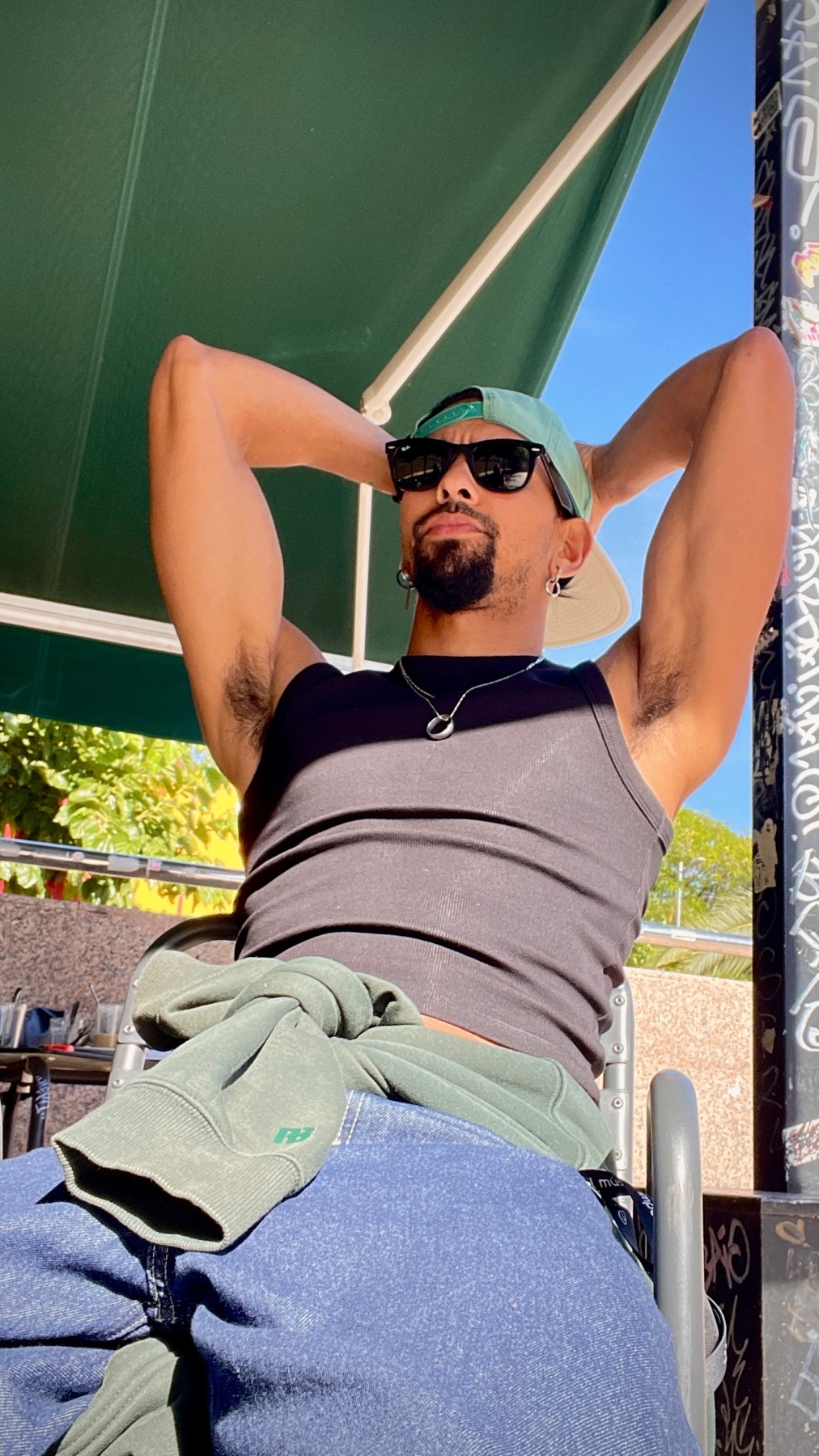 A man wearing sunglasses and a baseball cap poses outdoors with his arms behind his head. He is wearing a tank top and jeans.