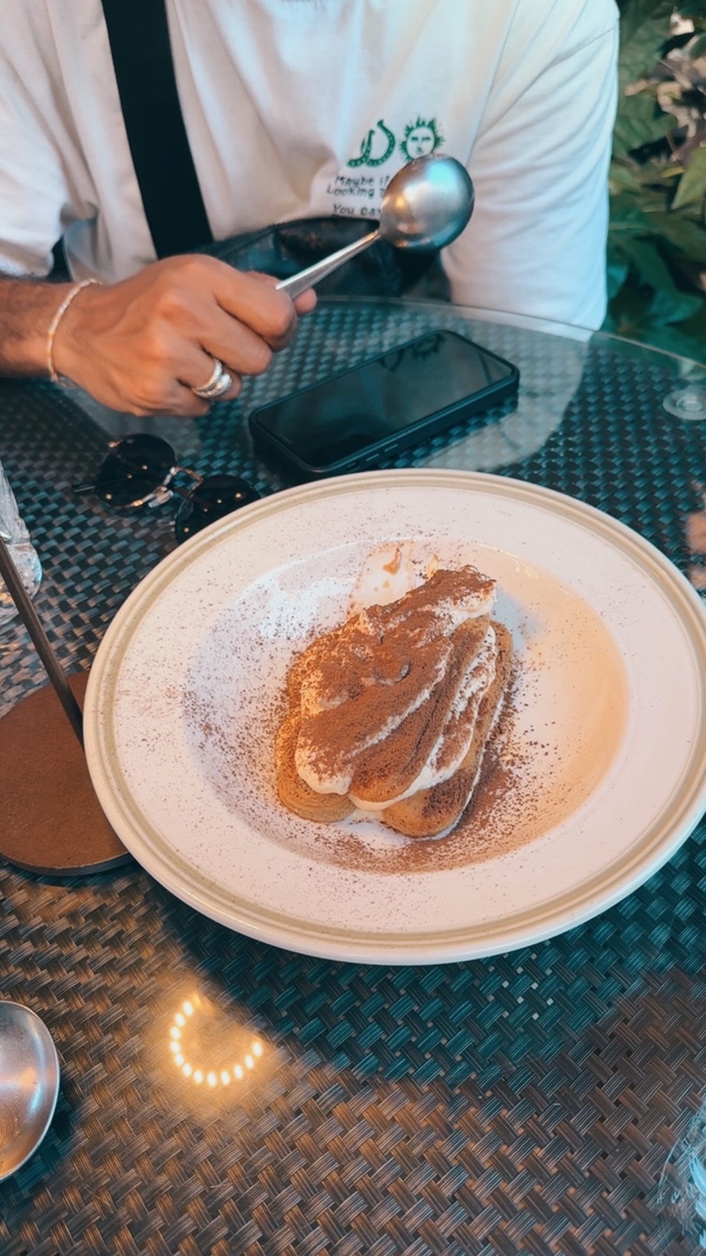 A close-up shot of a delicious tiramisu dessert on a white plate, dusted with cocoa powder. The dessert is ready to be eaten, with a spoon resting on the side of the plate.
