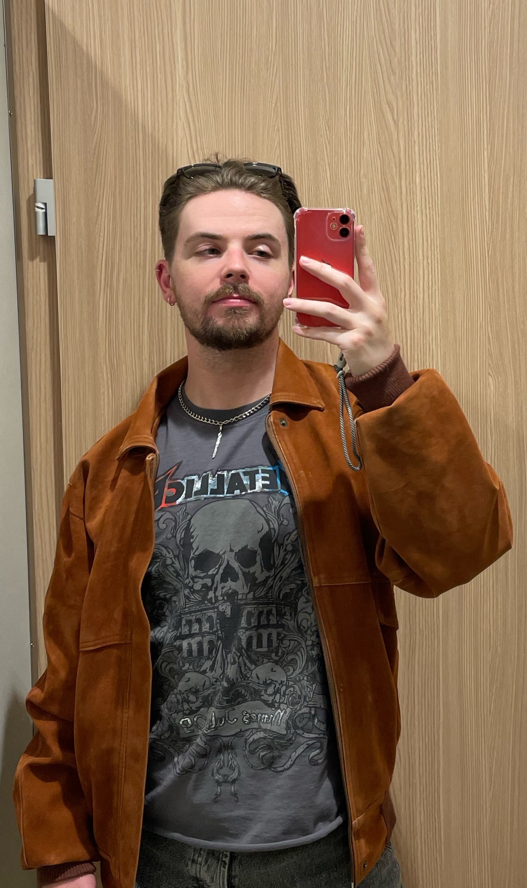 A man takes a mirror selfie, showcasing his outfit. He wears a brown jacket and a graphic t-shirt.