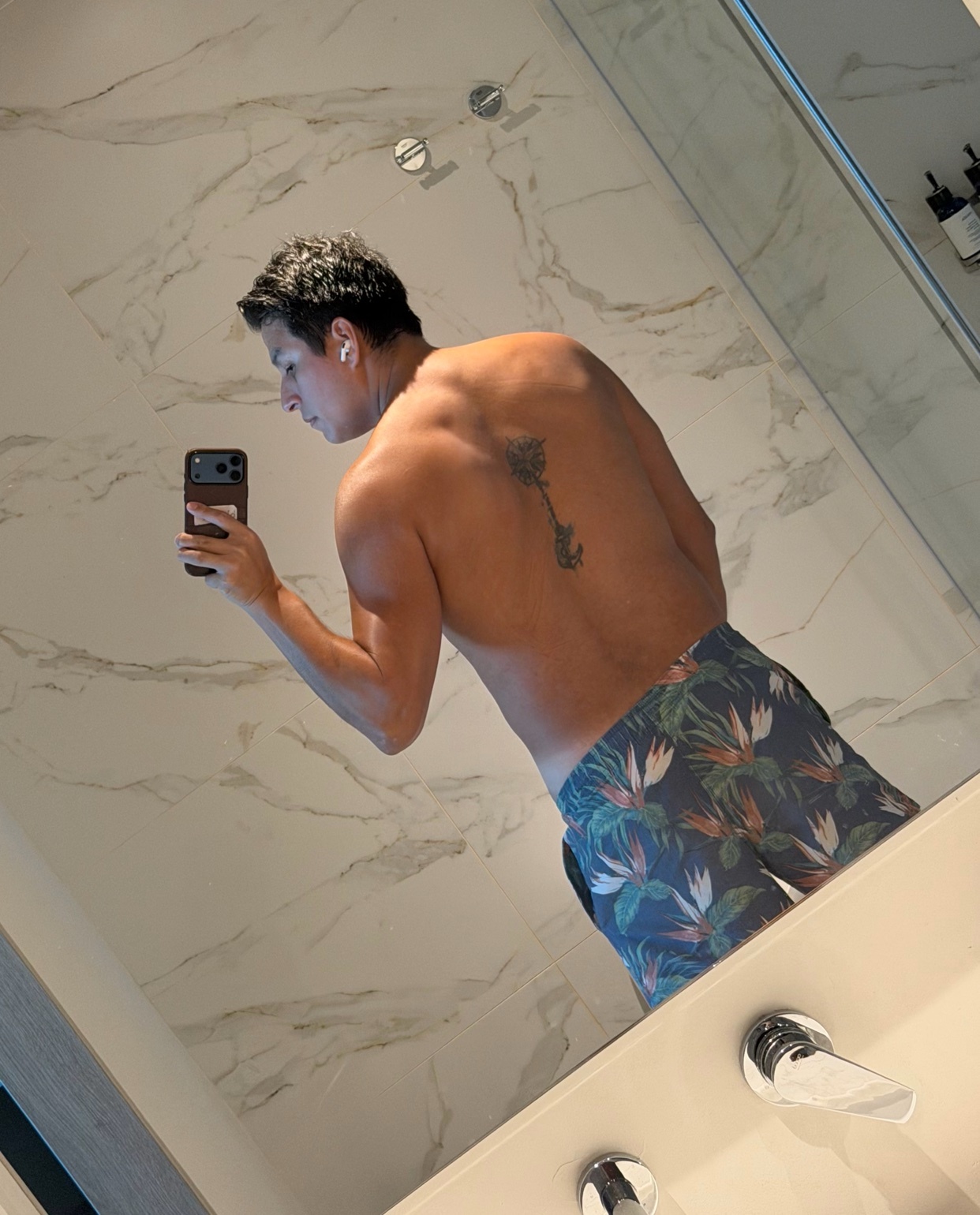 A man takes a mirror selfie in a bathroom, showing off his muscular back with a tattoo of a palm tree. He is wearing patterned swim trunks.