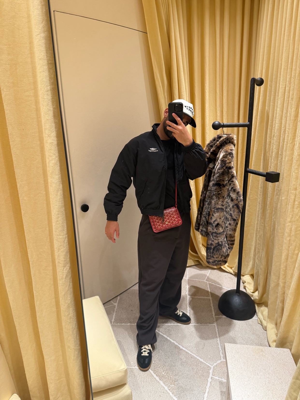 A man poses in front of a mirror, showcasing his outfit. He wears a black jacket, cap, and pants, with a red bag.