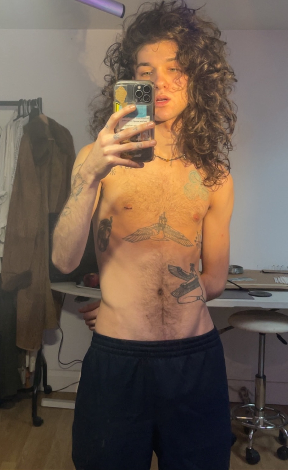 A shirtless person with long, curly hair takes a mirror selfie. They have tattoos on their chest and stomach.